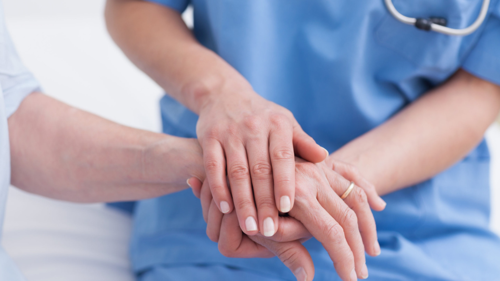 Nurse in blue scrubs, wearing a stethoscope holding the hand of a patient