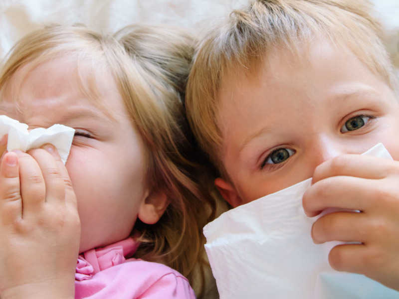 Image of two children lying in bed holding tissues to their nose
