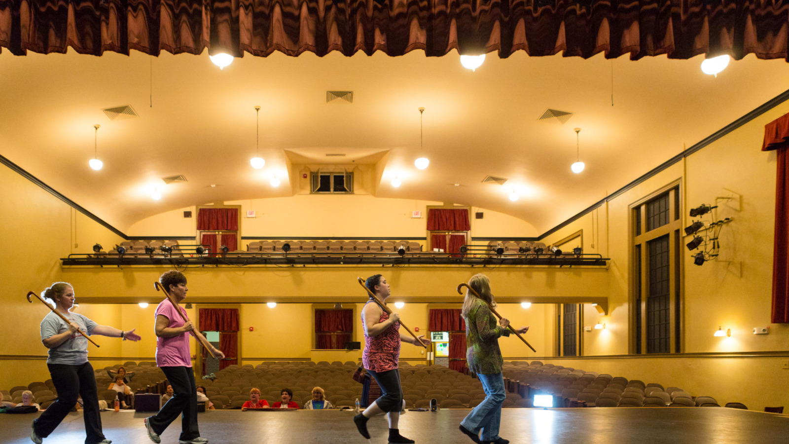 four performers walking across a stage with canes over their shoulders, whille a small audience looks on