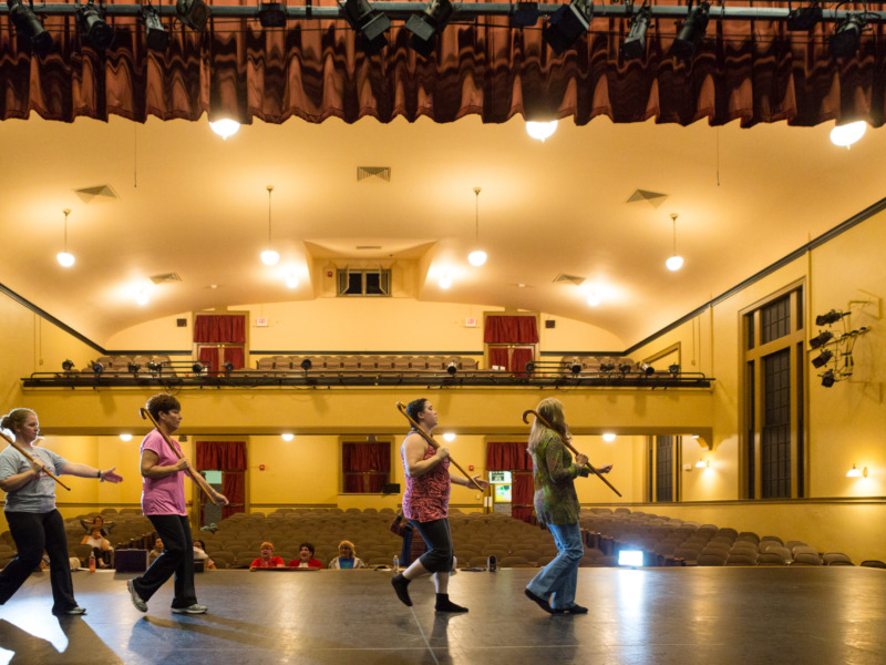 four performers walking across a stage with canes over their shoulders, whille a small audience looks on